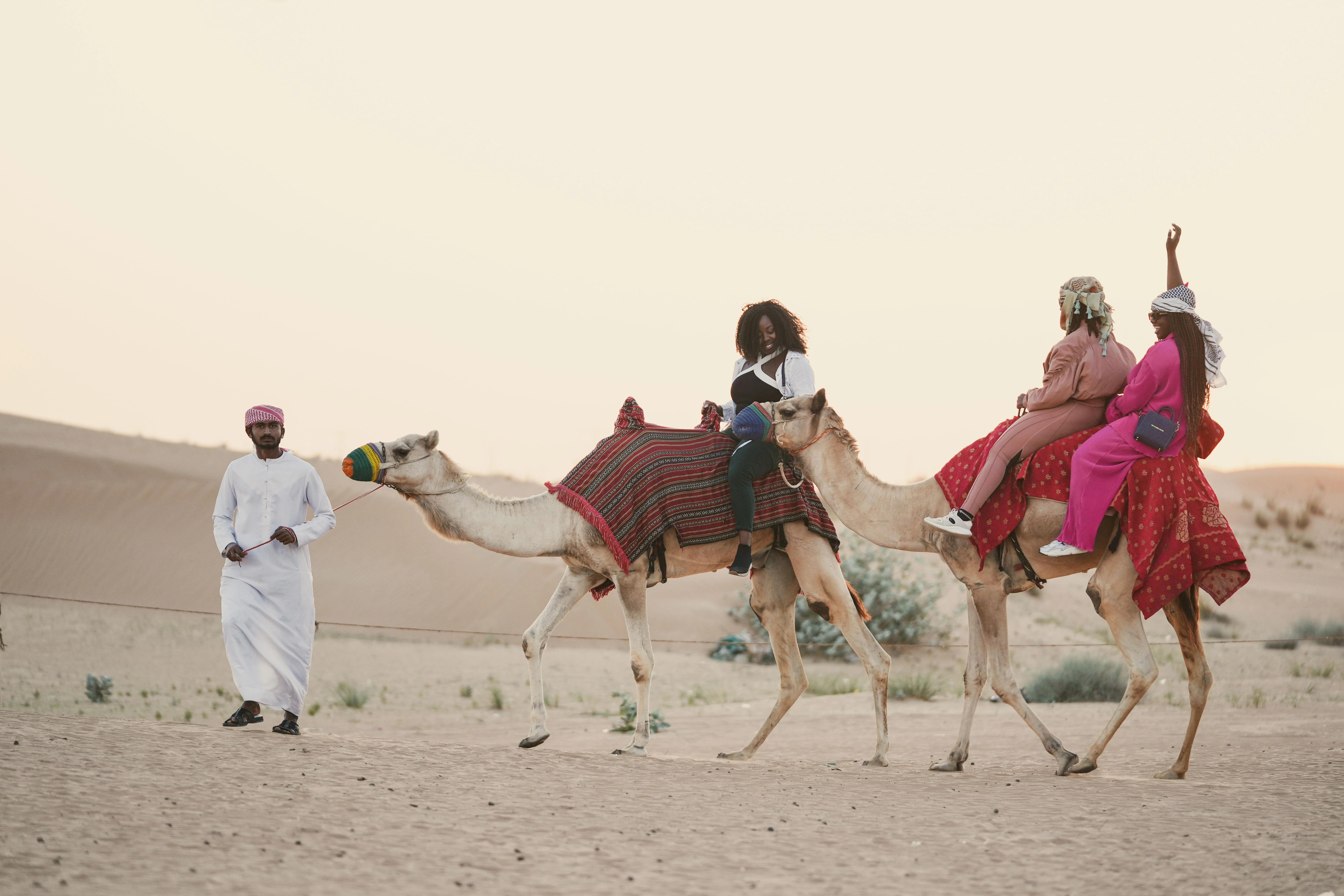 Guests riding camels through the desert with a guide walking alongside.