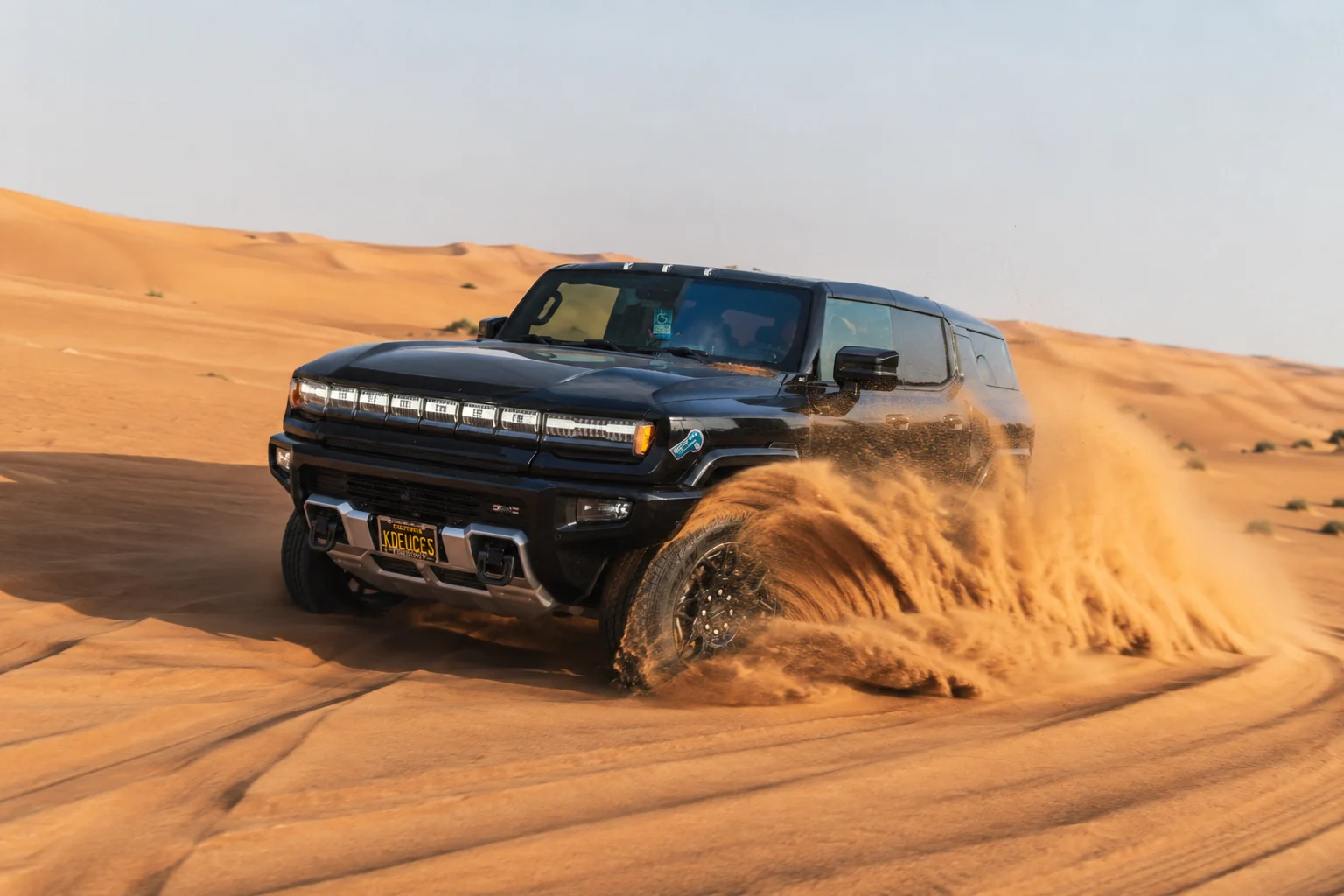 A black Hummer driving through desert dunes with sand spraying from the tires.